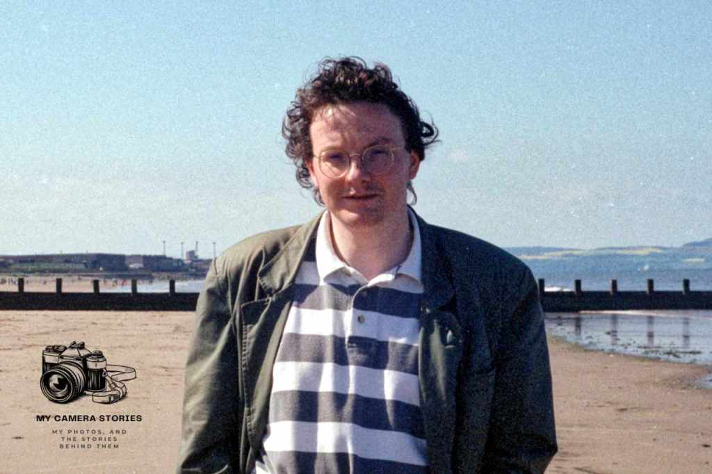 A person standing on Portobello Beach, wearing a striped shirt and jacket, with a neutral expression and a scenic background of the beach and sky.