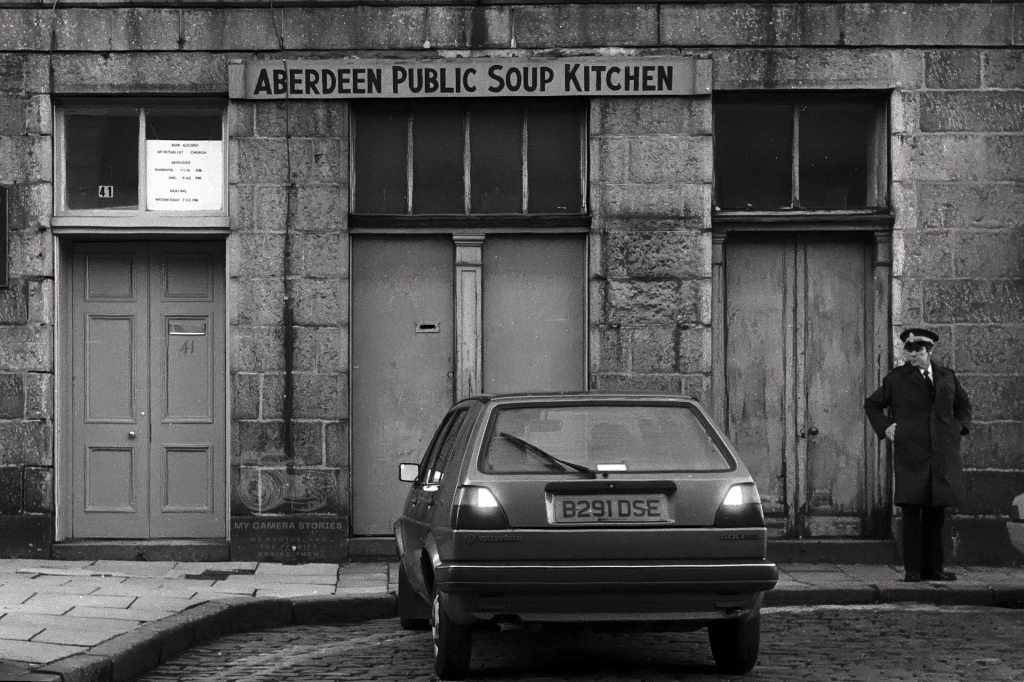Black and white photograph of the Aberdeen Public Soup Kitchen with a traffic warden standing nearby and a parked car in front.
