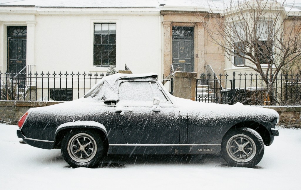 A vintage car covered in snow parked in front of a residential building on a snowy day.