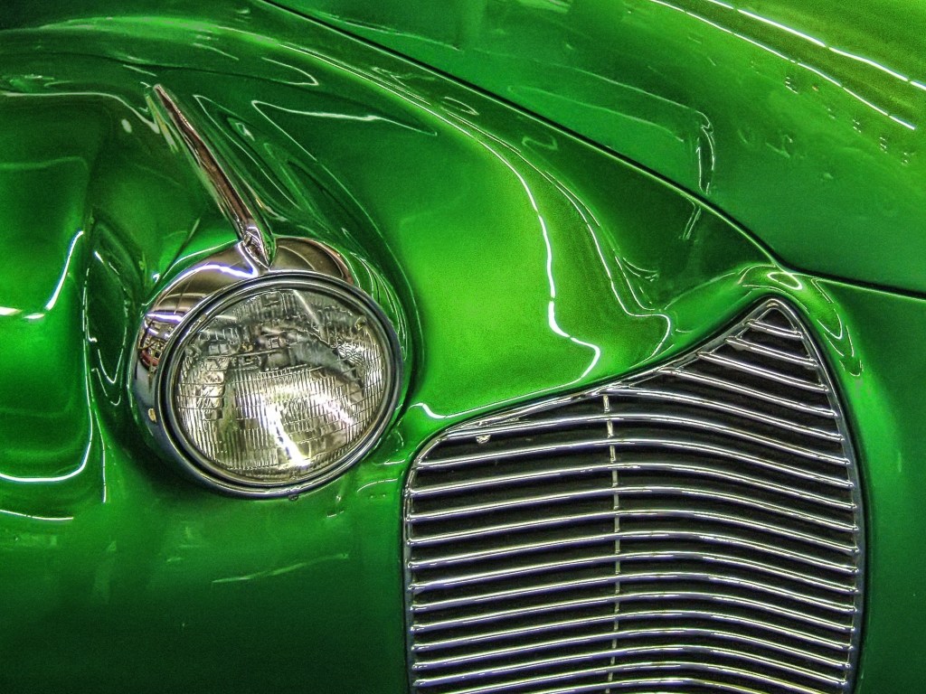 Close-up of a vintage car's green body with a round headlight and a chrome grille.