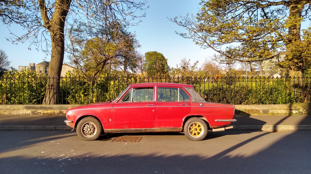 A vintage red car parked on the side of the street, surrounded by greenery and trees.