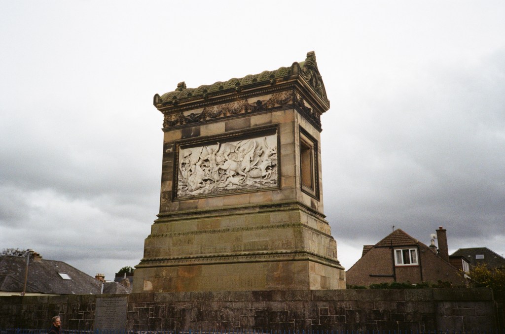 A stone monument with a sculpted relief on one side, situated in an urban setting under an overcast sky.