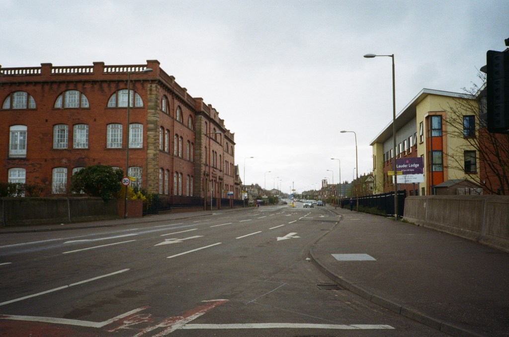 A street view in Scotland during the 2020 lockdown, showcasing deserted roads lined with red brick buildings and a modern structure. The overcast sky adds to the quiet atmosphere.