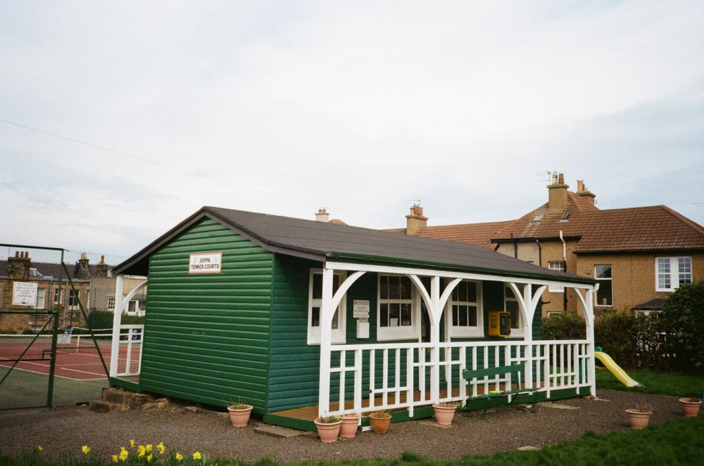 A green wooden building with white trim, labeled 'Joppa Tennis Courts,' alongside tennis courts in a residential area.