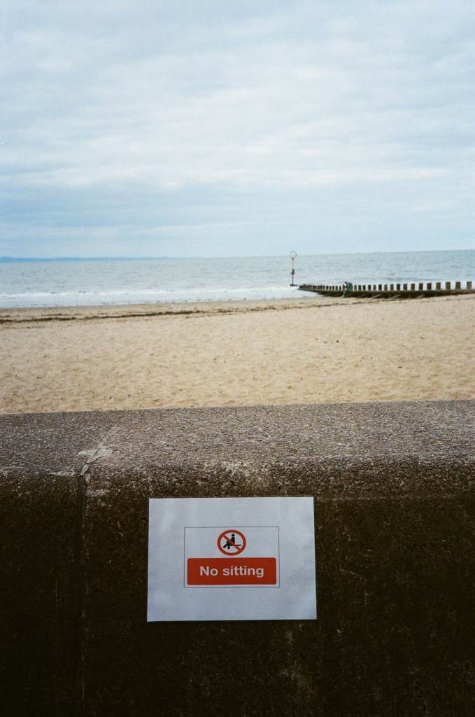 A 'No sitting' sign displayed on a concrete surface with a beach and sea in the background.