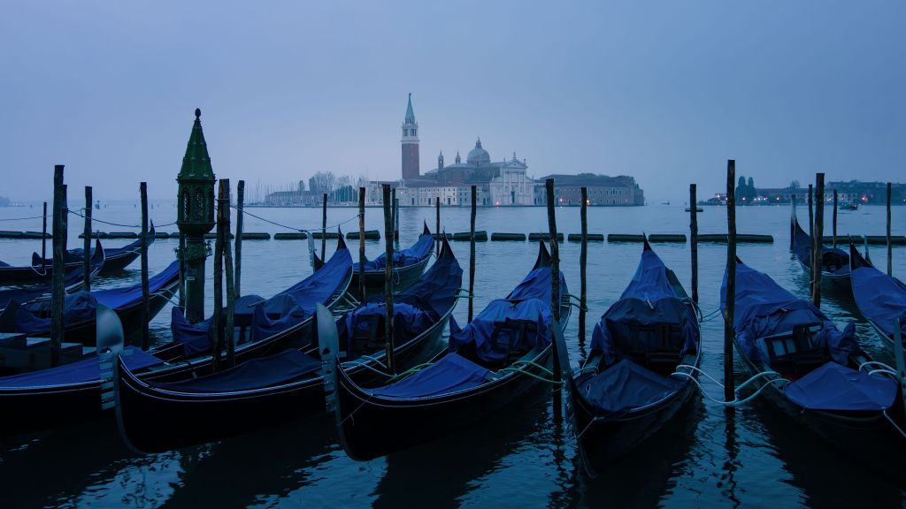 A tranquil scene of gondolas with blue covers moored along wooden posts in a calm lagoon, with a historic building and a tower visible in the background under a foggy sky.