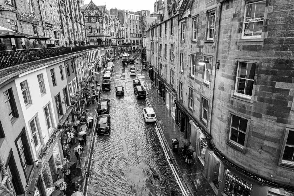 A high-angle view of Victoria Street in Edinburgh on a rainy day, with glistening cobblestones, parked cars, and people walking with umbrellas.