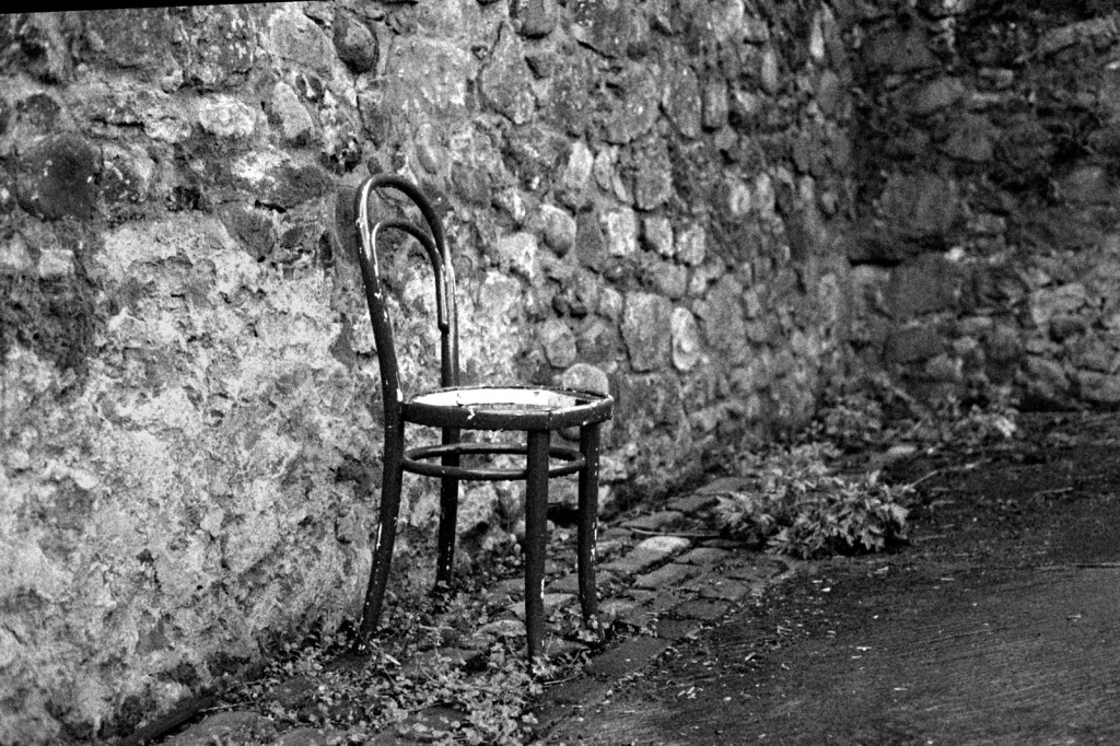 A solitary, weathered chair leaning against a stone wall, surrounded by cobblestones and vegetation, evoking a sense of abandonment and the passage of time.