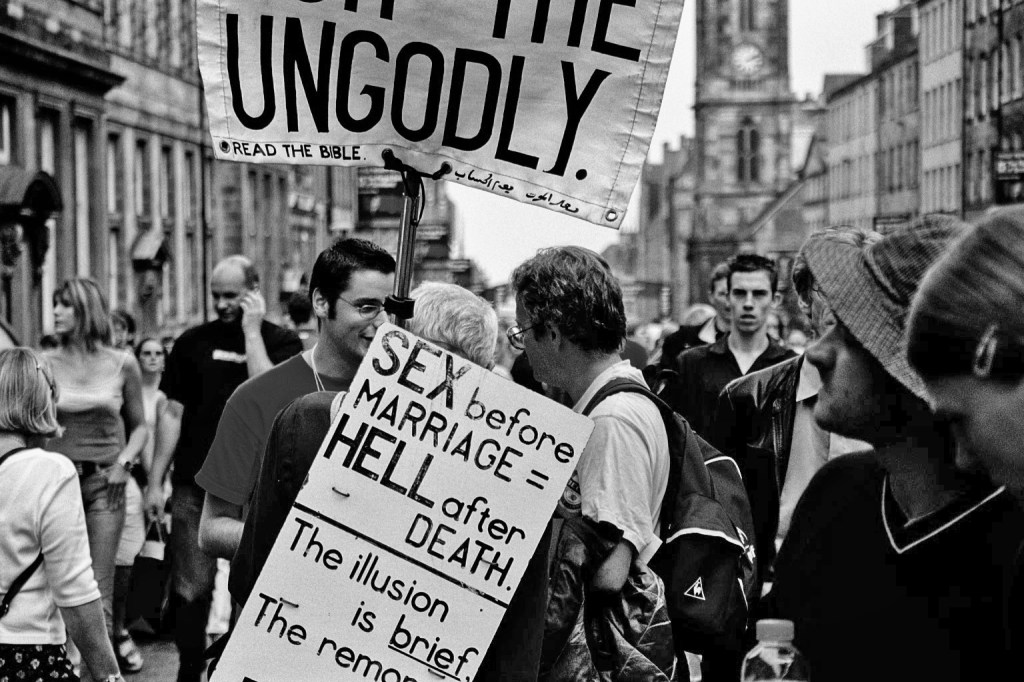 A preacher holds a placard with a warning about premarital sex on the Royal Mile during the Edinburgh Festival Fringe, engaging with a young man in front of a crowd. Surrounding them are various festival-goers, some glancing at the interaction while continuing with their activities.