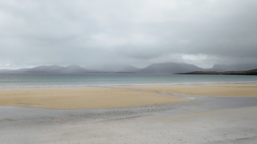 A tranquil view of Luskentyre Beach on the Isle of Harris, featuring soft sand and a calm sea beneath an overcast sky, with distant misty mountains in the background.