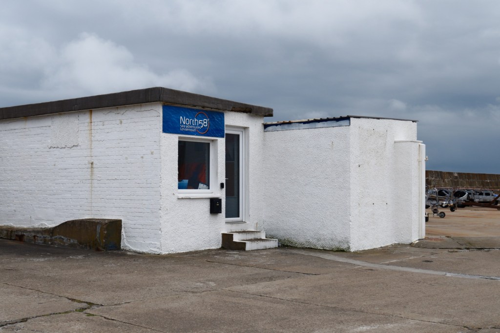 A small, white building with a blue sign reading 'North58' in Lossiemouth Harbour, against a cloudy sky.