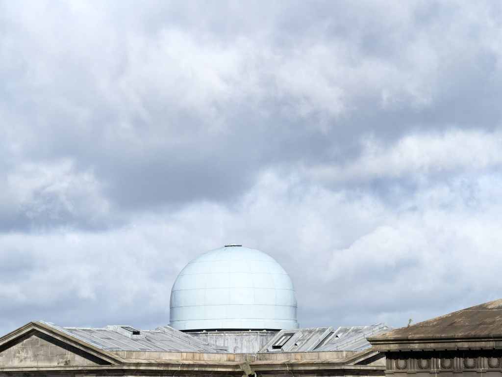 A cloudy sky above a dome-shaped structure, partially obscured by rooftops.