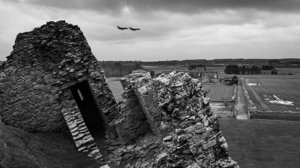 Black and white image of the ruins of Duffus Castle in Morayshire, Scotland, with blurred fighter jets flying in the sky above and farm buildings visible in the background.