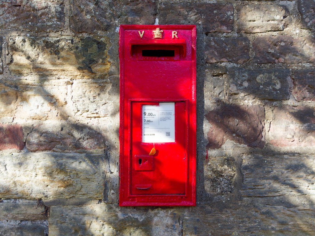 A red Victorian post box mounted on a stone wall, displaying a collection time notice.