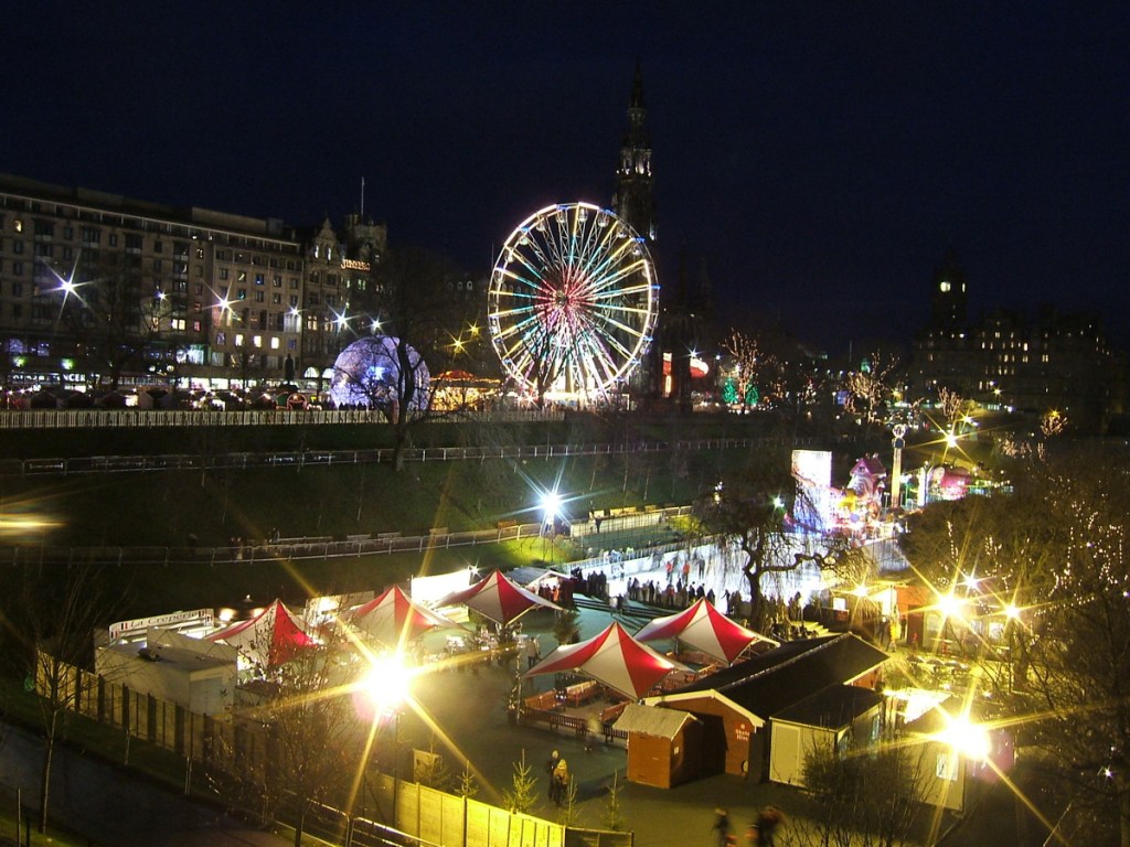 Nighttime view of a festive market with a large ferris wheel, brightly lit tents, and holiday decorations.