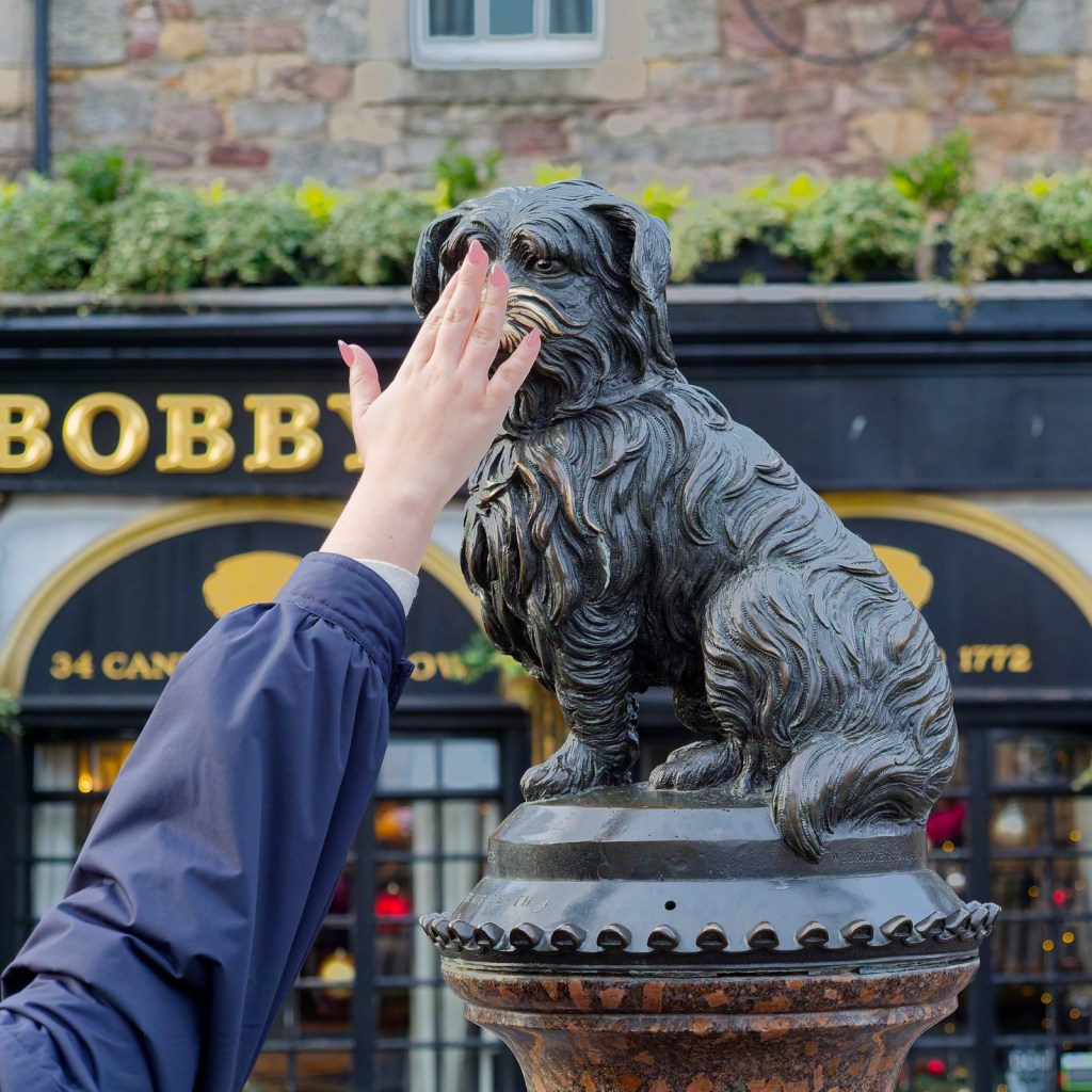 A person's hand reaching out to touch the nose of the Greyfriars Bobby statue, with a pub in the background.