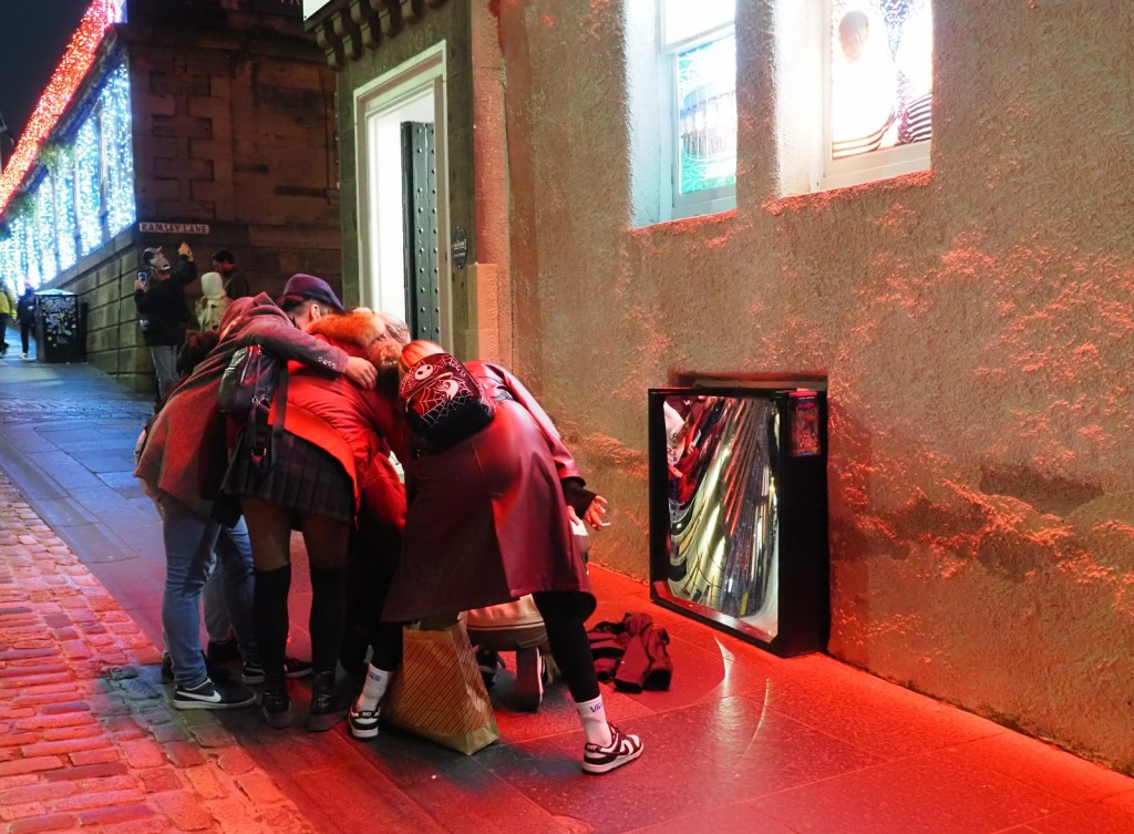 A group of people gathered around a distorted mirror on a street decorated with festive lights, creating a lively atmosphere.
