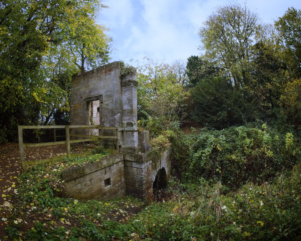 An old stone ruin surrounded by overgrown vegetation and trees, with a wooden fence in the foreground.