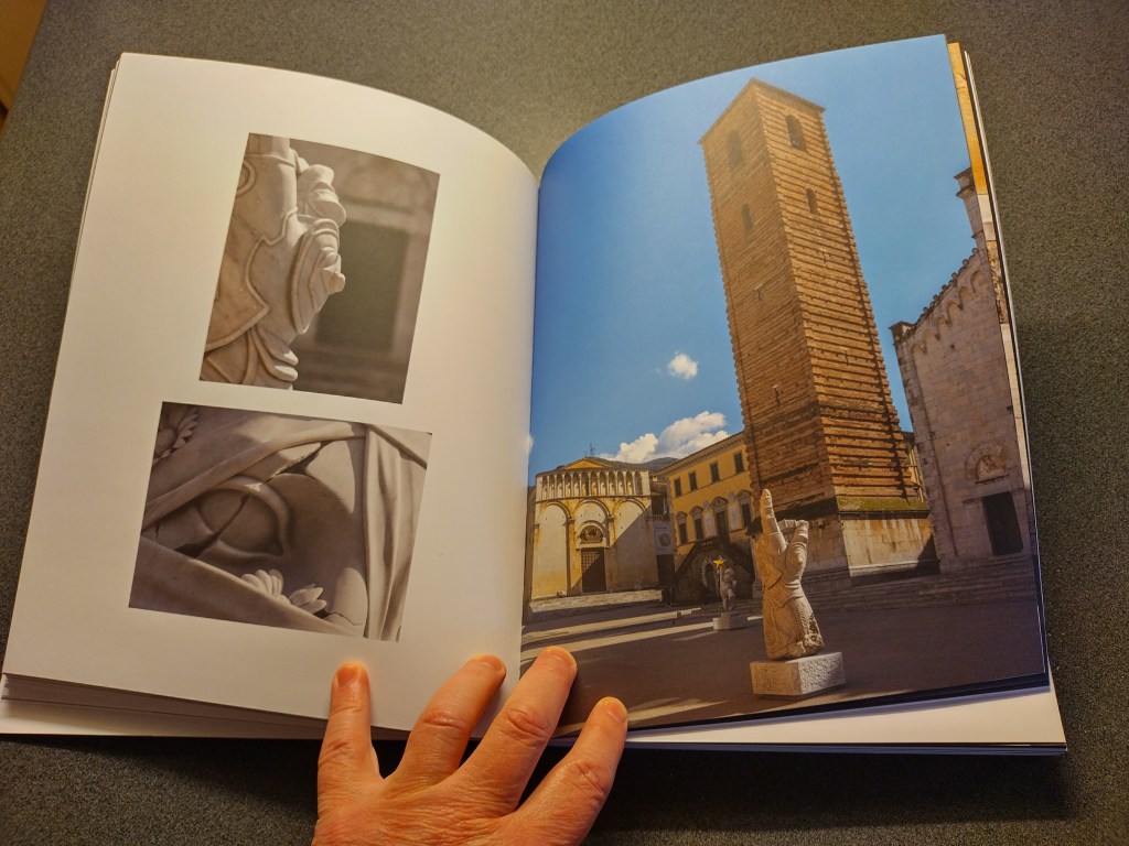 An open book featuring two photographs of intricate sculptures on the left page and a view of a tall, historic bell tower and buildings in a square on the right page. A hand is seen holding the book.