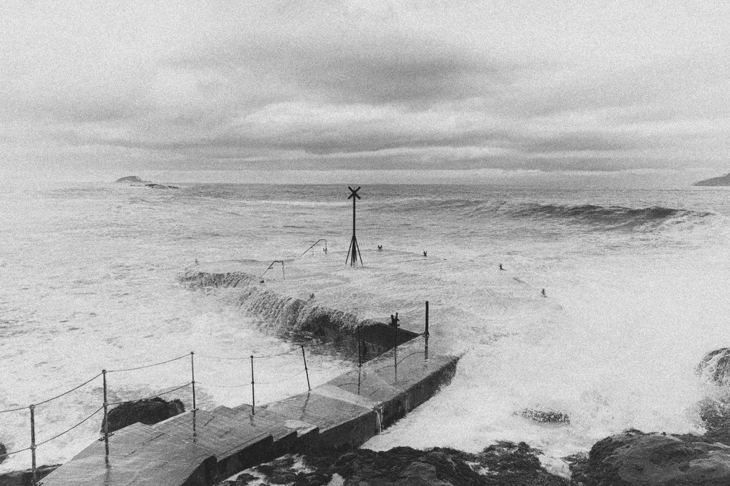 Black and white image of a rocky coastline with crashing waves, featuring a structure with a marker post and several surfers in the water.