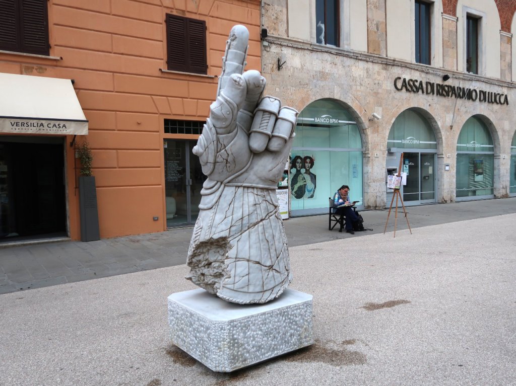 A large marble sculpture of a hand making a 'number one' gesture, set on a decorative base, with buildings and a seated person in the background.