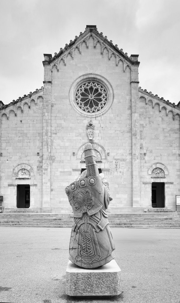 A close-up view of a large stone sculpture of a hand wearing an ornate gauntlet, set against a background of an old stone building with a circular rose window and intricate architectural details, in black and white.