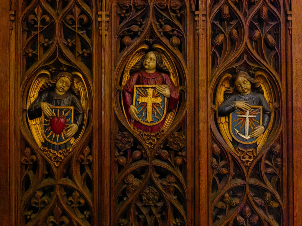 Intricate wooden carvings featuring three angels holding shields, adorned with symbols, against a backdrop of ornate foliage.